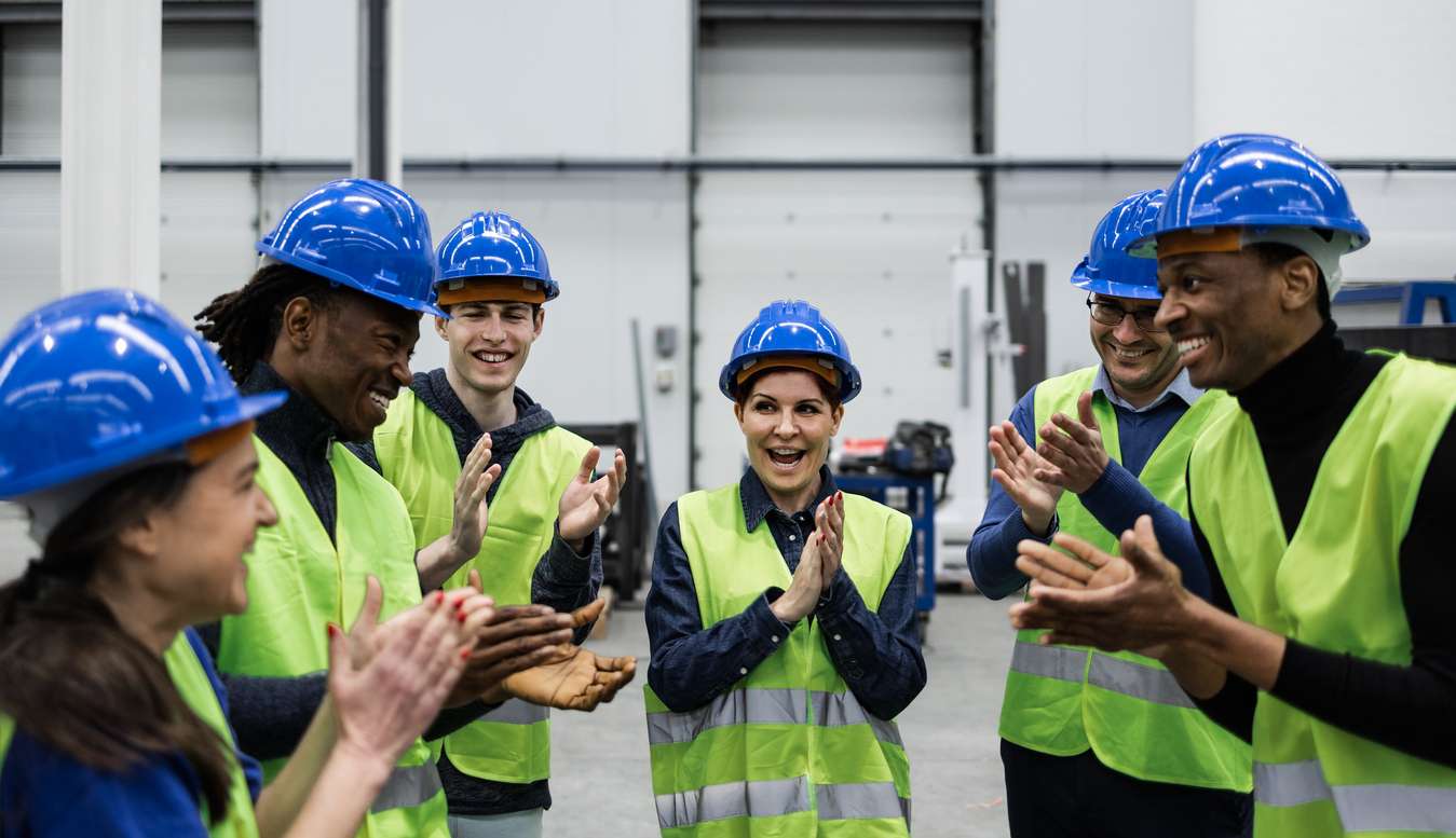 A semi circle of men and women in hard hats and safety vests smiling and clapping