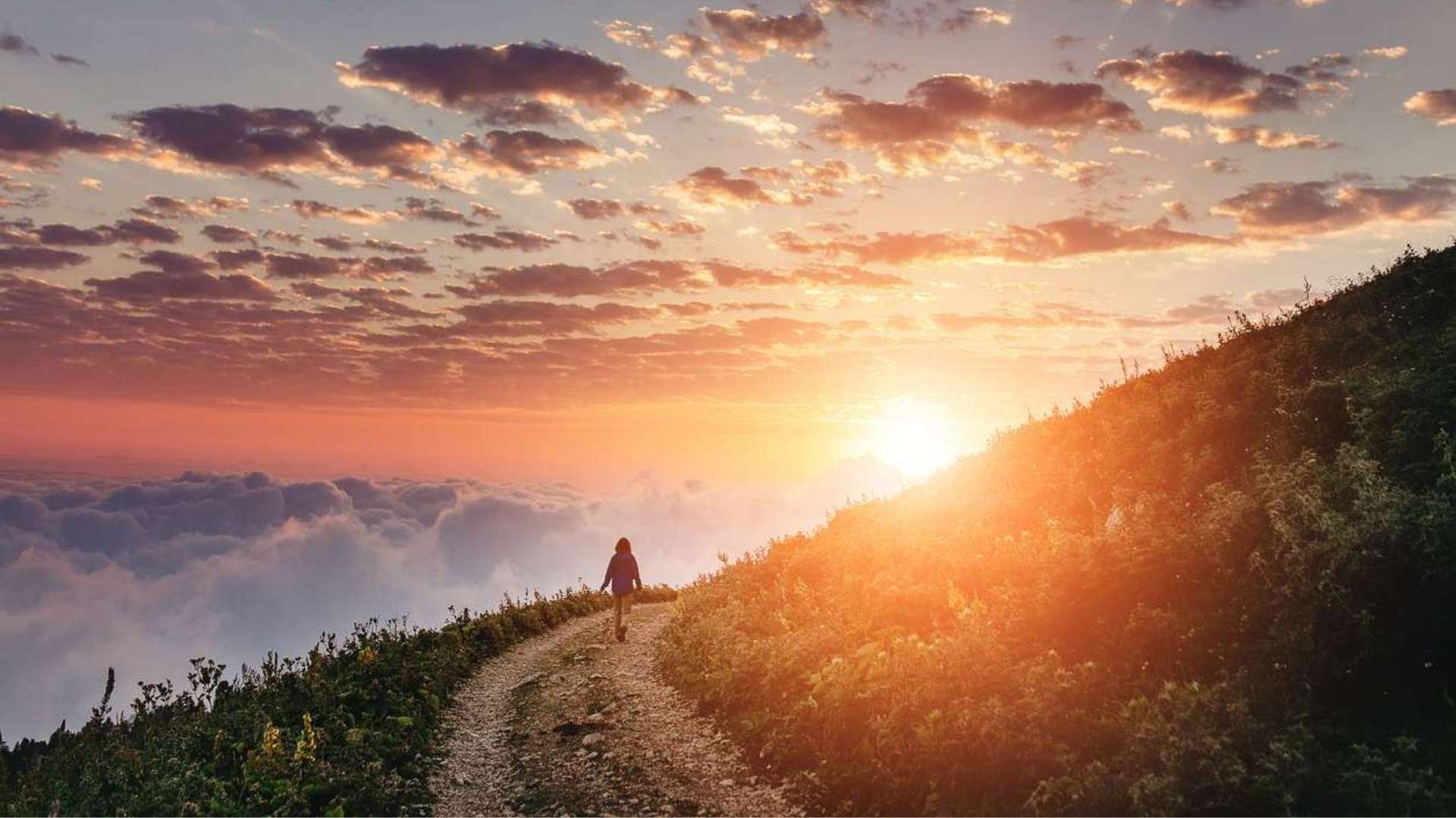 A vast image of the sun setting over a mountain, with a person walking beneath a cloudy sky at dusk.